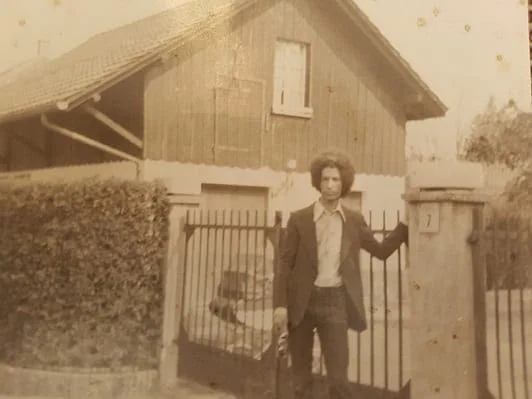El Arbi Bouqdib as a young man in front of a wooden house with a fence, old sepia photograph